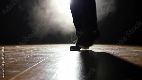 Legs in black pants and shoes tap dancing under a spotlight on a wood stage