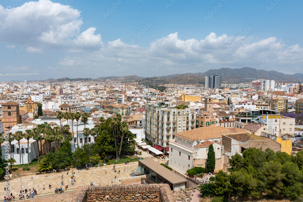 Obraz premium Elevated view from Alcazaba of Málaga’s cathedral and white city rooftops, framed by palm trees and clouds over the mountains.