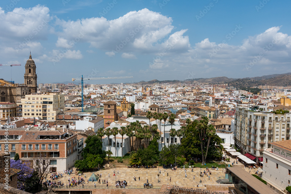 Obraz premium Elevated view from Alcazaba of Málaga’s cathedral and white city rooftops, framed by palm trees and clouds over the mountains.