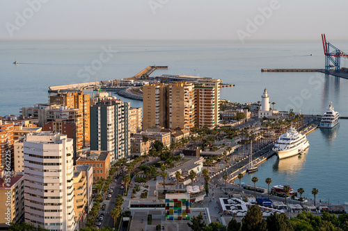 Iconic lighthouse and vibrant seafront of Málaga at sunset, with luxury yachts, palm-lined boulevards, and urban life by the sea.