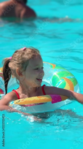 Happy little girl laughs and plays in blue hotel pool with rubber ring. Child fun swims and splashes with inflatable circle. Summer vacation holiday travel. Vertical shot
