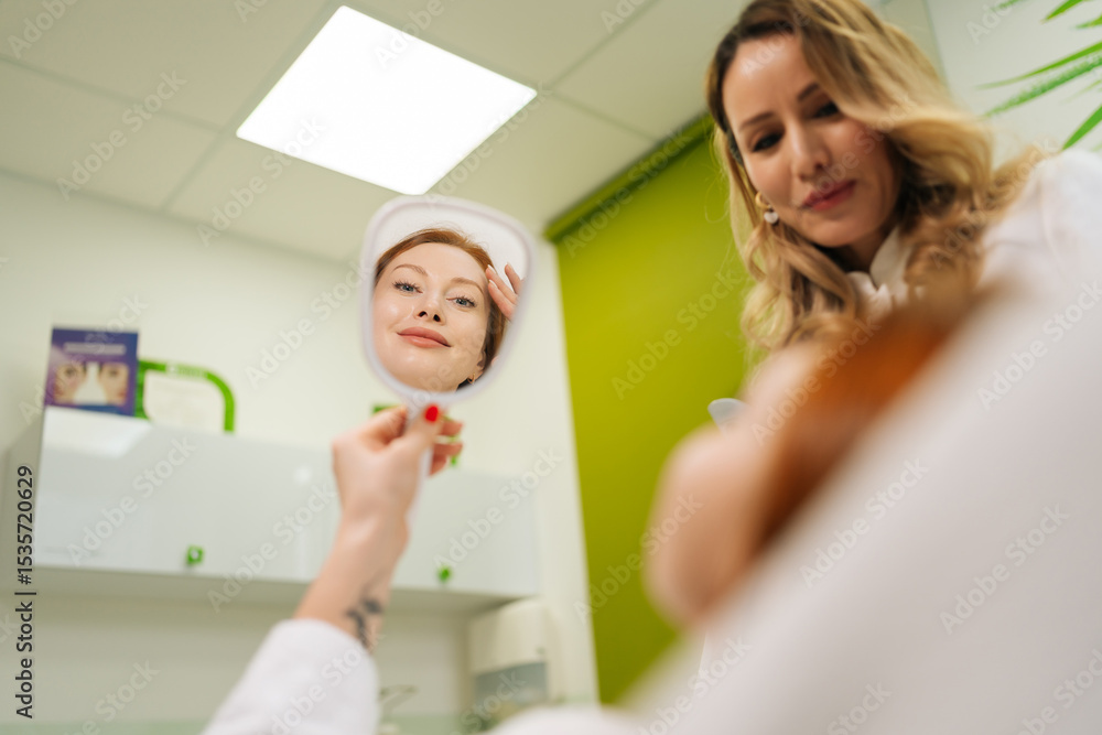 Fototapeta premium Happy young woman admiring reflection in mirror, showcasing radiant skin after successful beauty treatment, while beautician smiling, pleased with impressive results. Concept of beauty treatment.