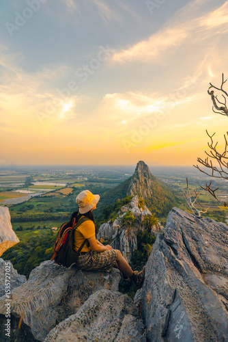 Picture from the back of a woman sitting on the peak of a high mountain cliff. There are a lot of rock with white color. The sun is setting on the mountain and there is a beautiful warm orange light.
