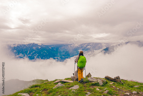 Photo from the back of a female hiker standing at a high mountain pass surrounded by dramatic clouds and rocky alpine terrain. Idea for a solo trekking adventure in the Himalayas.