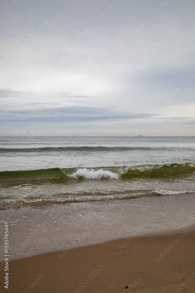 White foamy waves in a beautiful blue sea. In the distance, silhouettes of ships can be seen sailing near the coast of Gdansk on the Baltic Sea. Wave after wave rolled onto the shore. Background. White foamy waves in a beautiful blue sea. In the distance, silhouettes of ships can be seen sailing near the coast of Gdansk on the Baltic Sea. Wave after wave rolled onto the shore. Background.