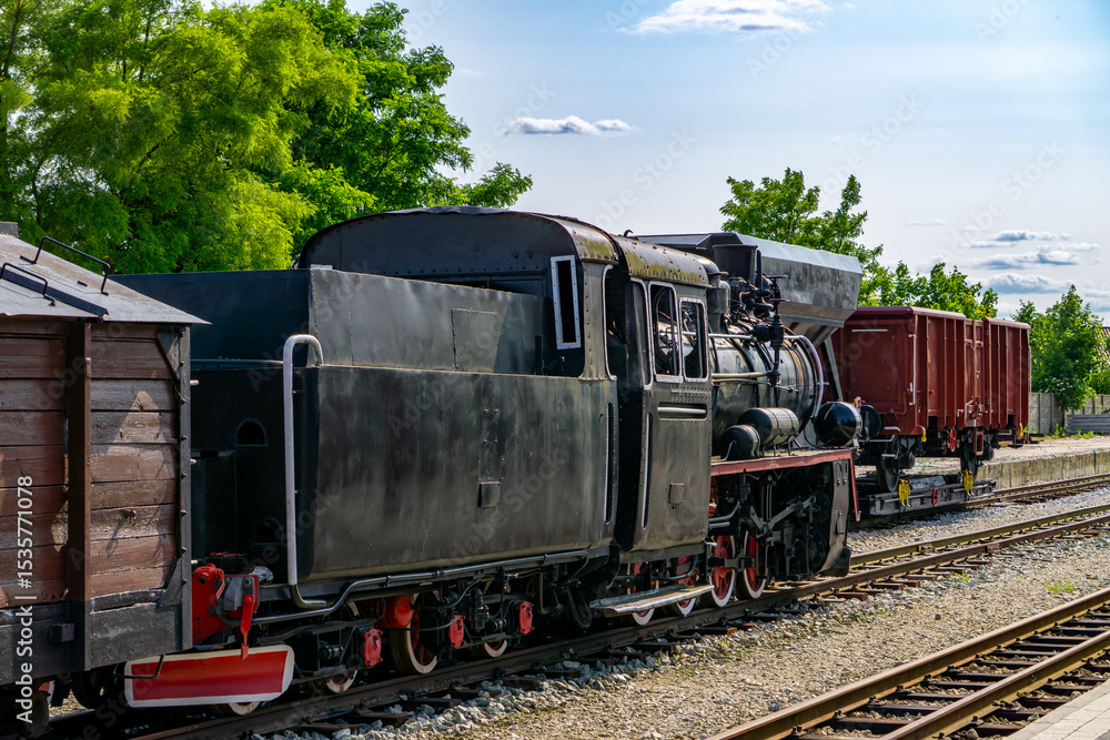 Obraz premium old, historic narrow-gauge steam locomotive with freight cars at the narrow-gauge railway station in Zbiersk, Greater Poland Voivodeship, surrounded by greenery on a sunny day.