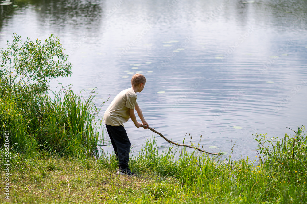 Boy standing on lakeside with a long stick, playing near the water in nature on a summer day Boy standing on lakeside with a long stick, playing near the water in nature on a summer day