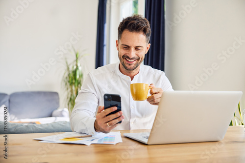 Smiling freelancer using mobile phone at home while working on laptop