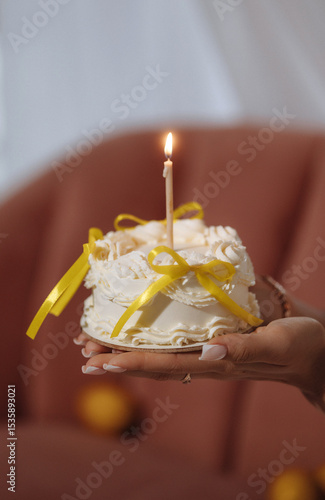 Wallpaper Mural Female hands with long manicured nails holding small round cake with cream, yellow ribbons, and burning candle. Shot indoors in warm light. Concept: celebration, birthday, sweetness, femininity. Torontodigital.ca
