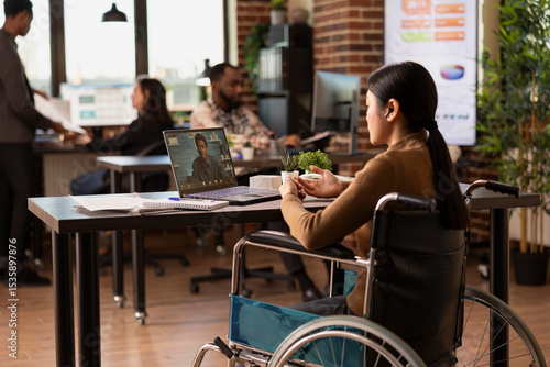Valokuva Female manager with a disability sitting in modern workspace, conducting a virtual meeting with a remote coworker
