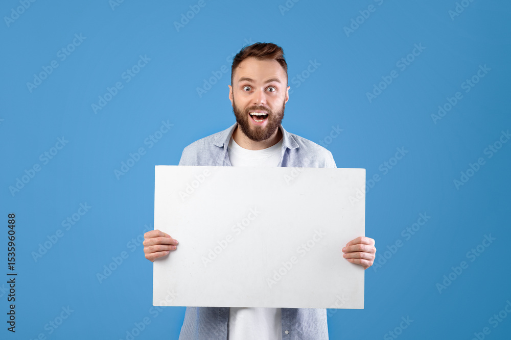 Excited young man holding blank poster with copy space on blue studio background. Happy millennial guy showing empty banner with mockup for your advertisement design Excited young man holding blank poster with copy space on blue studio background. Happy millennial guy showing empty banner with mockup for your advertisement design