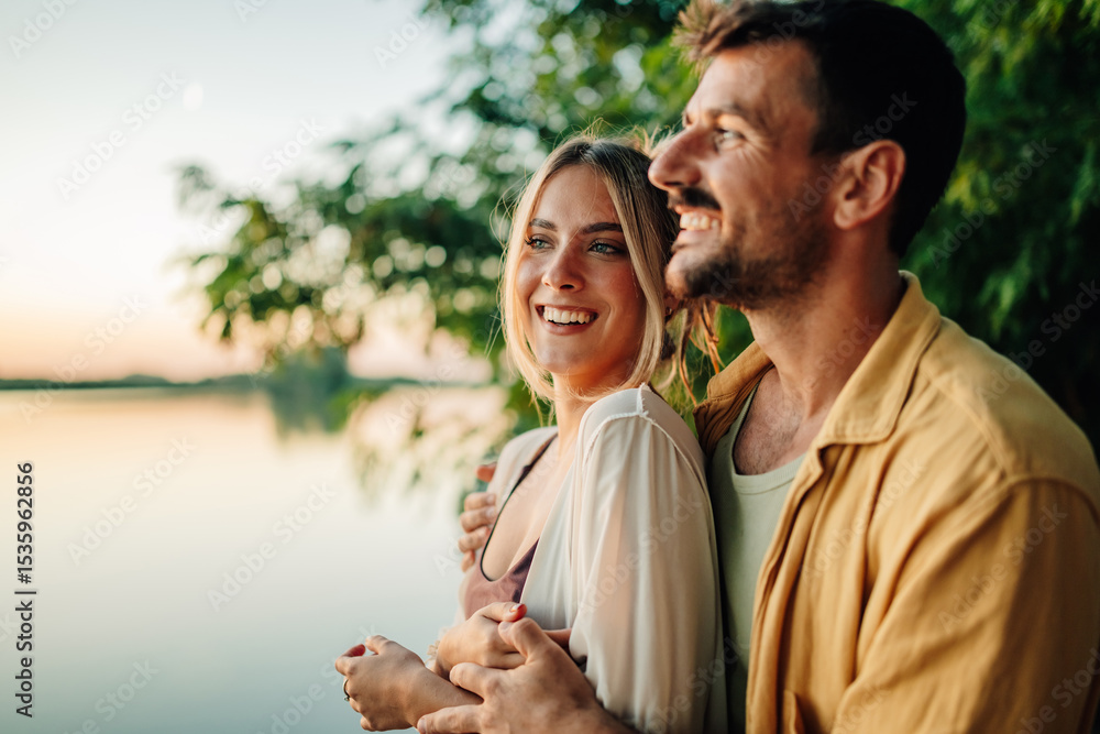 Fototapeta premium Happy couple embracing and smiling by the lake at sunset