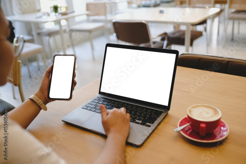 A person multitasking with a laptop and a smartphone, both with blank white screens. Ideal responsive design mockup for apps or websites in a cafe scene.