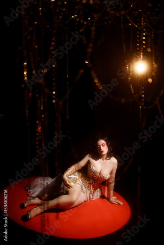 Wallpaper Mural Young Caucasian woman in ornate golden corset, lying on red round stage floor, under hanging beads, dark background, front view. Concepts: cabaret, circus, retro, burlesque. Torontodigital.ca