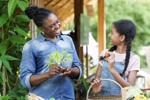 african woman and multiracial teenage girl holding vegetables and egg while smiling at outdoor organic farm showing joy and success of farming education in family-style agricultural environment