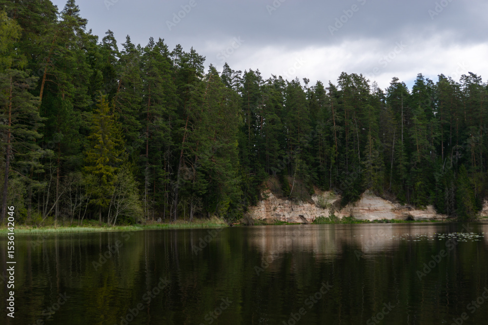 Fototapeta premium Mossy Cliffside with Dark Cave Entrance and Reflections in Calm Forest River Surrounded by Evergreen Trees