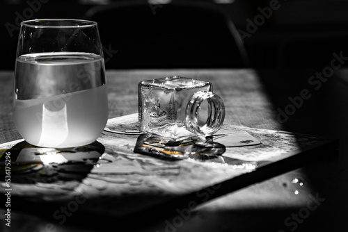 Glass of water and small square glass jar placed on a magazine in dramatic lighting, casting intricate shadows.