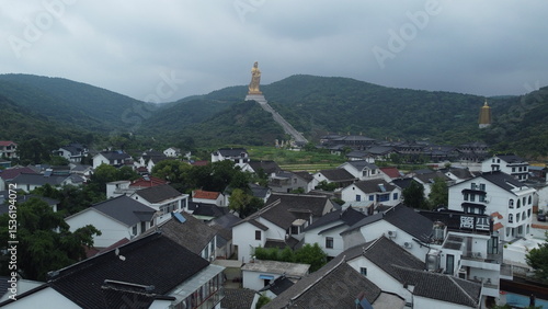 Aerial views of the golden Guanyin statue and temple complex near Wuxi, China. Surrounded by green hills and traditional village rooftops under cloudy skies
