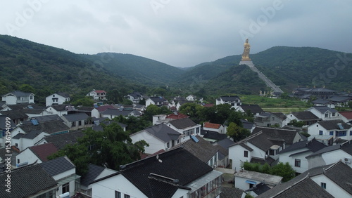 Aerial views of the golden Guanyin statue and temple complex near Wuxi, China. Surrounded by green hills and traditional village rooftops under cloudy skies