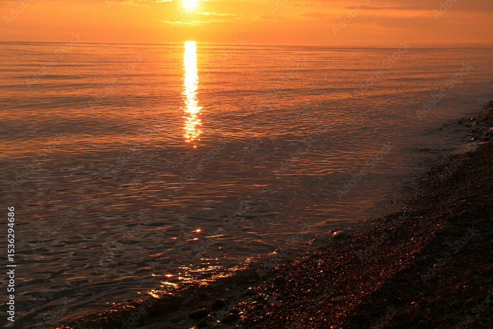 Obraz premium Soft waves gently lap pebbled beach under warm sunset glow. Horizon blurs in vibrant hues, creating serene coastal scene. Calm atmosphere captures natures beauty Batumi Georgia Adjara region