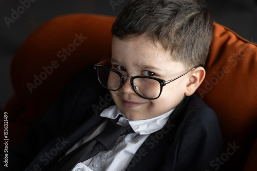 Slika na platnu Smiling kid dressed in business jacket with glasses lying on sofa, using laptop and talking on smartphone