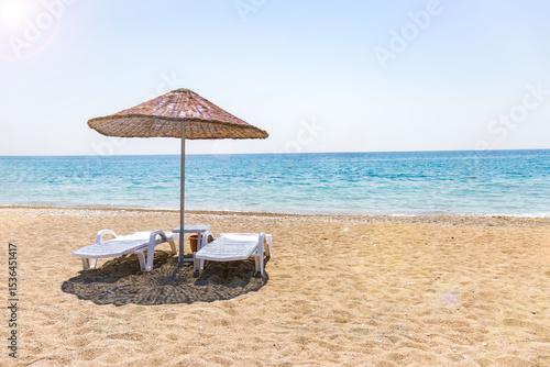 Two white sun loungers under a straw parasol on a sandy beach with turquoise sea and clear blue sky. Idyllic summer vacation concept.