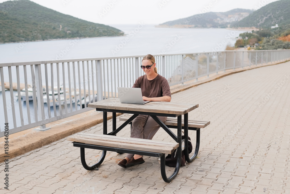 Fototapeta premium Young woman working remotely by the sea with a laptop and solar panel, embracing sustainable travel, mindful lifestyle and digital freedom. Concept of new work, balance and conscious living.