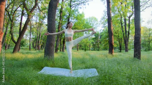 Reaching balance at golden hour, a woman stands poised on one leg in a yoga stretch. Amid tall grass in a quiet forest park, her focused posture reflects calm strength and flowing presence.