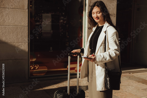 Close up of young woman with suitcase holding phone and getting ready for travel, standing in front of entrance to train station in Lisbon. Concept of business travel and individual tourism