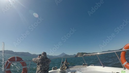 A tourist stands on a boat and takes pictures of nature on his phone. The boat is sailing on the water in the sea or ocean. Rocks or mountains are visible on the horizon.