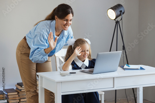 Online school education concept. Mother and child greet teacher online and wave at laptop screen. Schoolgirl learns e-learning lesson.