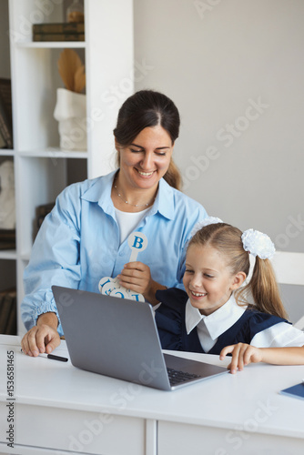 Vertical lifestyle portrait young mother sits at the table with daughter and helps little schoolgirl do her online homework on the computer at home. Woman tutor teaches a girl lessons on a laptop
