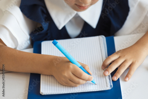 Closeup schoolgirl in a school uniform writes an assignment in a notebook with a blue pen during class lesson.
