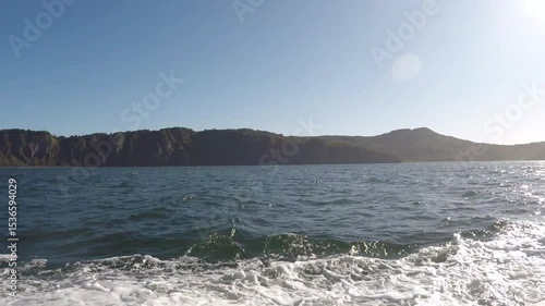 A boat is floating on water in the sea or ocean or lake. It is rocking on the waves. The sun is shining. Rocks or mountains are visible on the horizon. A view from the back of the boat.
