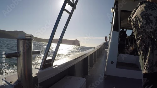 A boat is floating on water in the sea or ocean or lake. It is rocking on the waves. The sun is shining. Rocks or mountains are visible on the horizon