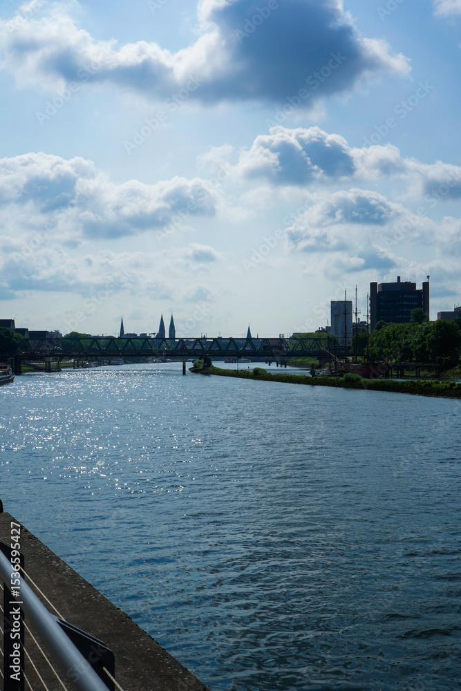 Obraz premium A calm river flowing under a bridge under a cloudy sky with the city skyline in the background. A large river through the city of Bremen, Weser in 28199 Bremen, Germany