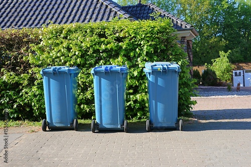 Three full blue bins with paper waste are standing by the hedge at the roadside ready for collection and emptying