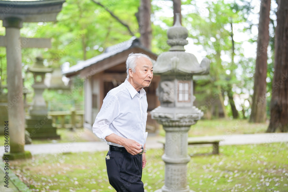 Fototapeta premium In rural Japan, a Japanese man in his late 70s visits a countryside shrine surrounded by lush green trees. Wearing a white shirt and hat, he prays quietly in the peaceful warmth of early summer air.