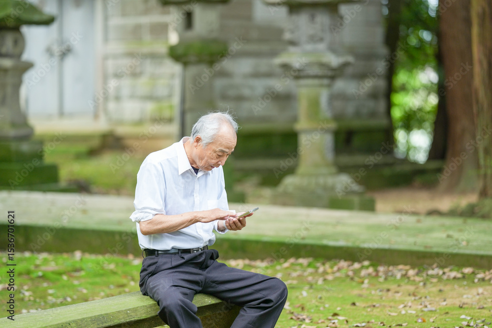 Fototapeta premium In rural Japan, a Japanese man in his late 70s sits at a peaceful countryside shrine, wearing a white shirt and hat. He uses his smartphone, surrounded by lush greenery in the early summer sunlight.