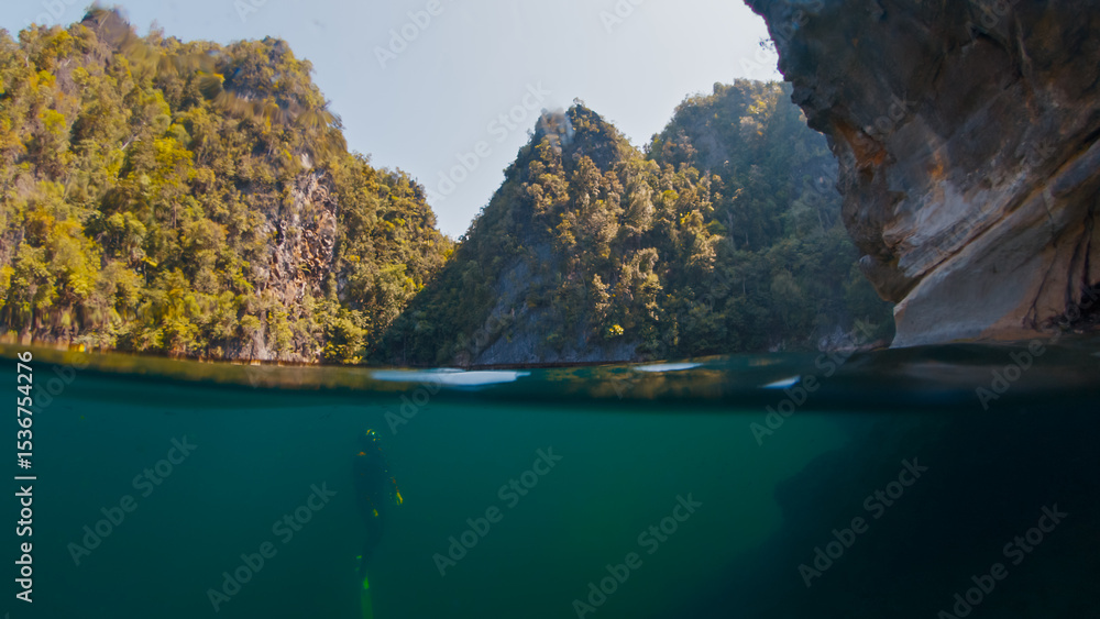 Obraz premium Freediver swims underwater in the closed lake. Splitted above and underwater view of the diver swimming in the lake surrounded by limestone mountains. West Papua, Misool, Indonesia