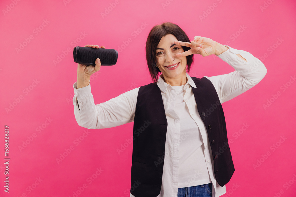 Businesswoman showing victory sign and holding wireless speaker on pink background Businesswoman showing victory sign and holding wireless speaker on pink background