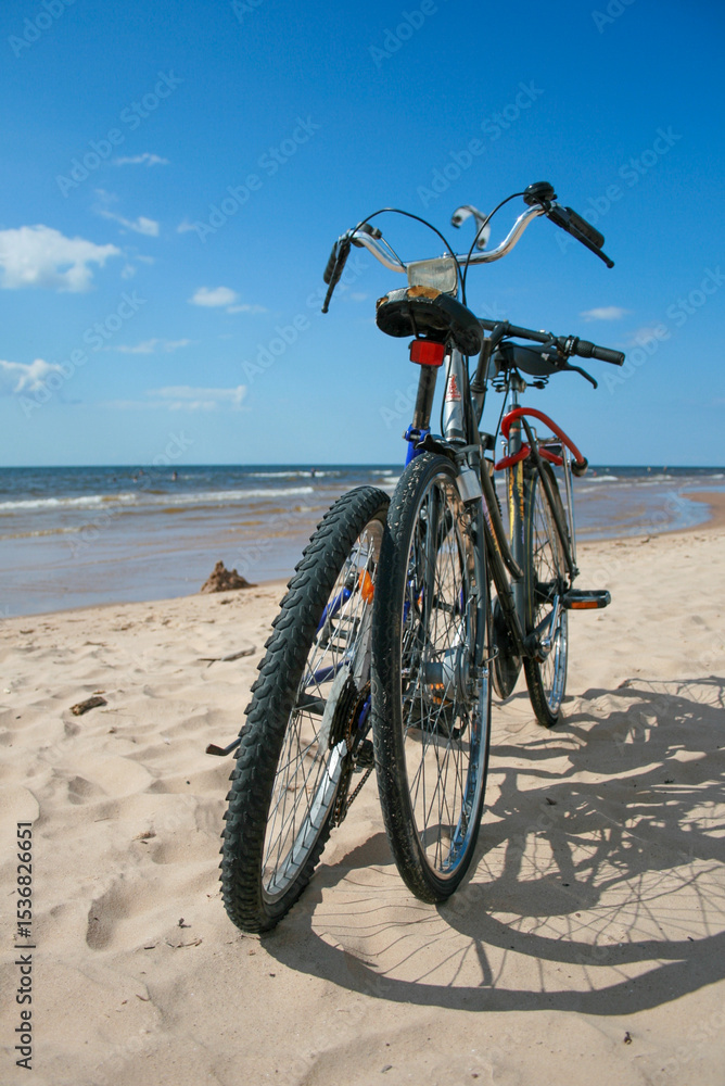 Fototapeta premium Two bicycles parked on a sandy beach with the sea and clear blue sky. Cycling to the beach on a bike for vacation leisure activity by the water. Swimming, sunbathing and relaxing