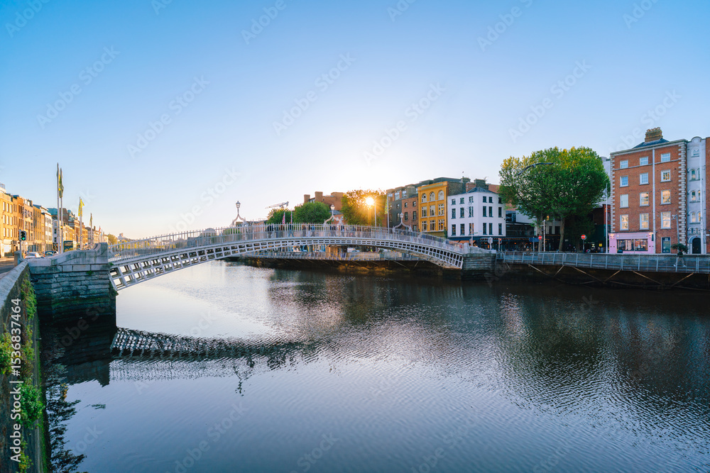 Obraz premium A view of Dublin’s Ha’penny Bridge over the River Liffey, framed by historic city buildings and a bright sky.