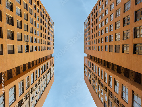 Striking upward view of two symmetrical modern high-rise buildings with orange facades, showcasing urban architecture, geometry, and city living under a clear blue sky.