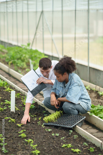 Male scientist wearing white lab coat pointing to soil while Young African female researcher holding plant seedling tray in greenhouse
