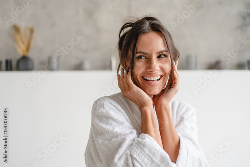 Beautiful young Caucasian brunette lady in bathrobe enjoying sunny morning in luxury bathroom touching her face fresh glowy skin, looking aside away on copy space. Wellness beauty routine concept