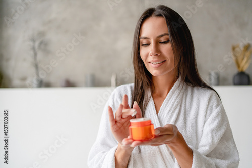 Photos Portrait of a young beautiful Caucasian woman in bathrobe smiling applying face body cream on her face for rejuvenation soft moisturizing effect
