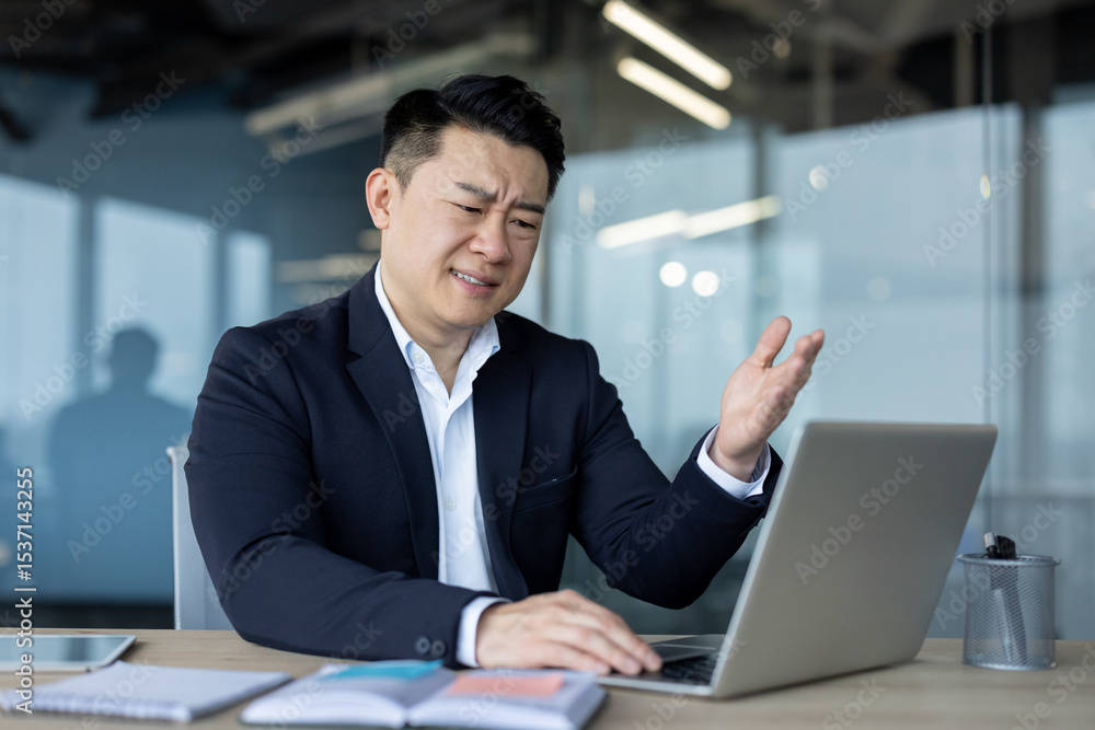 Dissatisfied young Asian male businessman sitting at desk in office and looking at laptop gesturing with hand Dissatisfied young Asian male businessman sitting at desk in office and looking at laptop gesturing with hand