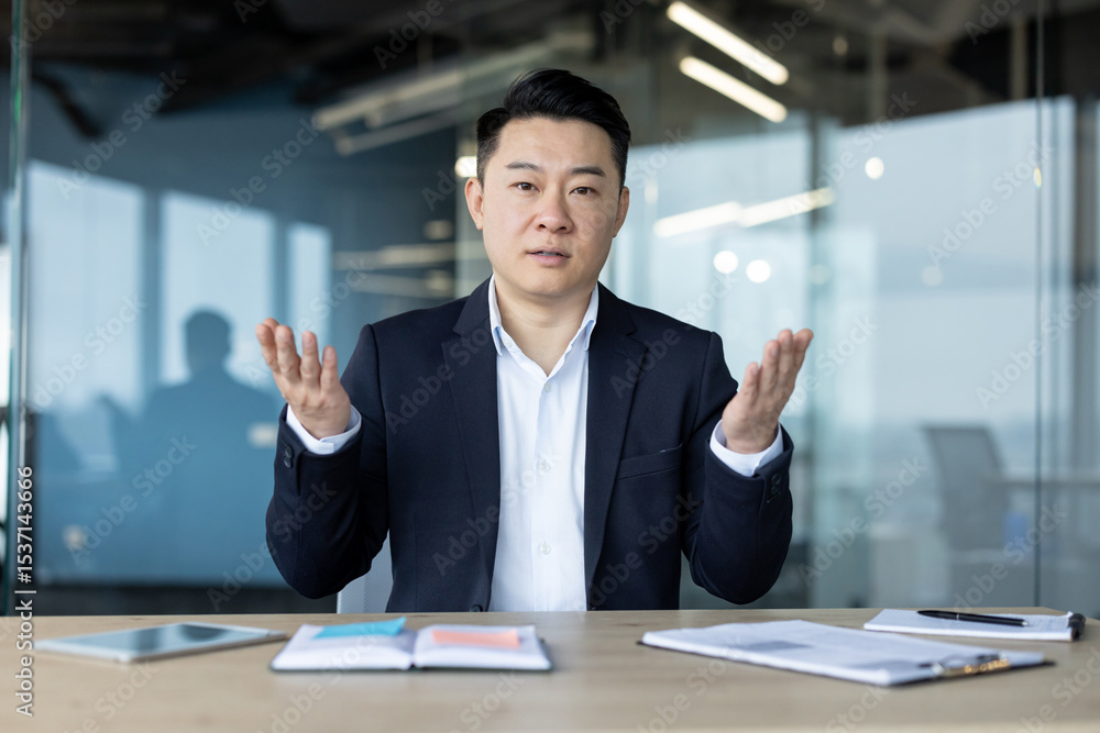 Portrait of a young Asian businessman in a suit sitting at a desk, talking and explaining to the camera Portrait of a young Asian businessman in a suit sitting at a desk, talking and explaining to the camera