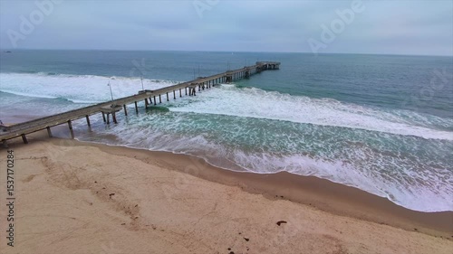 Vista aérea de muelle en la playa con olas y cielo gris.